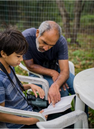Young boy with binoculars looking in a book with a man pointing at a page