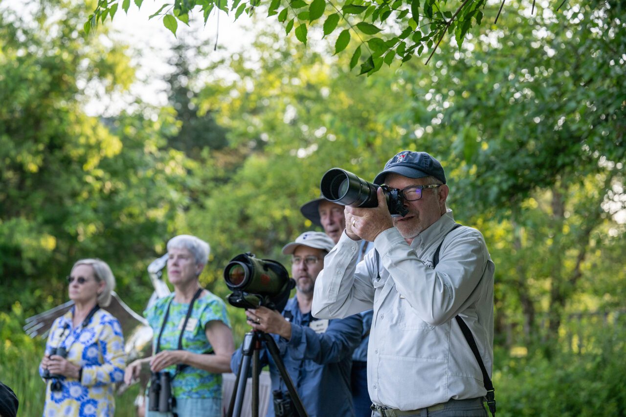 A group of birdwatchers outdoors. A man in the foreground looks through a camera with a long lens, while others stand behind him with binoculars and a spotting scope. Green trees surround them.