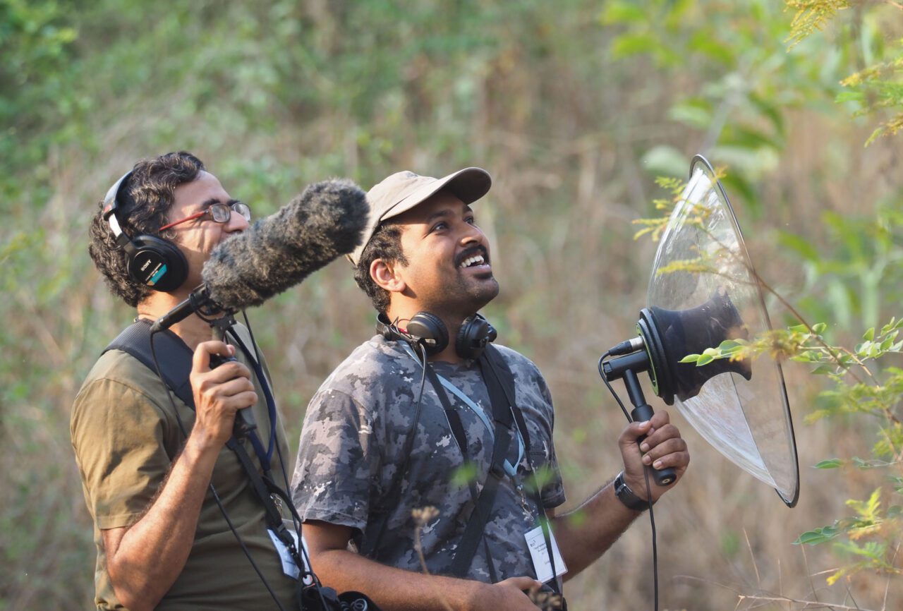 Two men recording bird sounds in the field. One holds a large microphone with a windscreen, while the other uses a parabolic microphone.