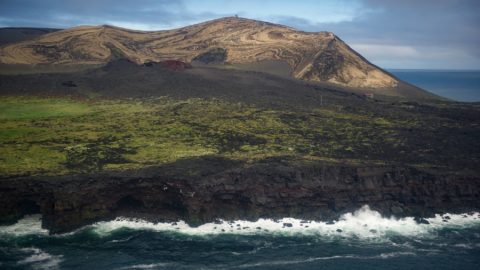 a volcanic island with a grassy meadow among lava plains, with ocean waves crashing against cliffs
