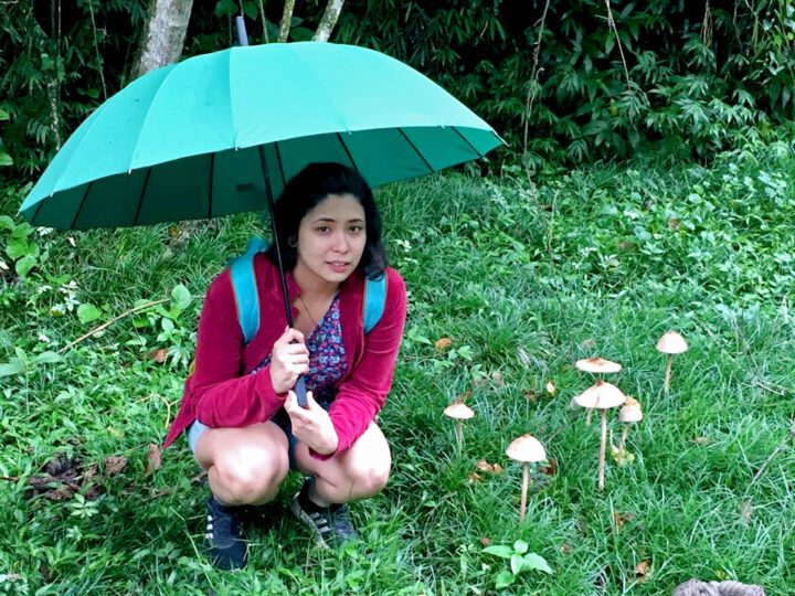 woman squats under a hand-held umbrella next to some large mushrooms in the grass.