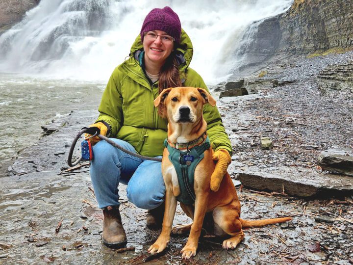 Woman with dog in front of a waterfall, wrapped up against the cold and rain.