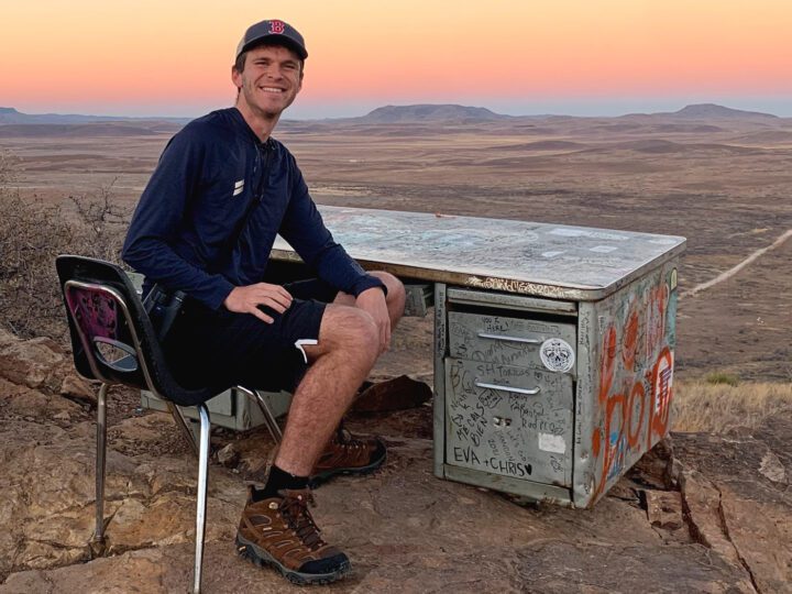 A man sits at a desk outside with a sunset behind him. Appears to be in a desert.