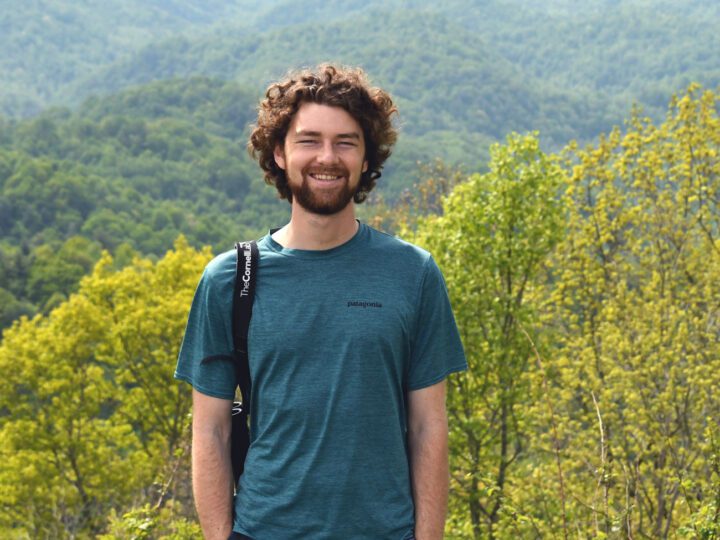 Bearded man in green shirt stands on forested mountain vista.