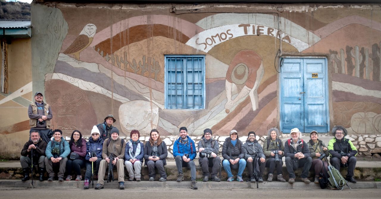 A group of people with cameras and binoculars sit against a wall with a mural.