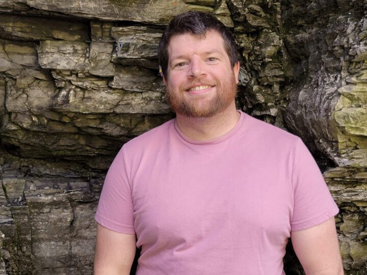 Man in a pink shirt stands against a rock wall.