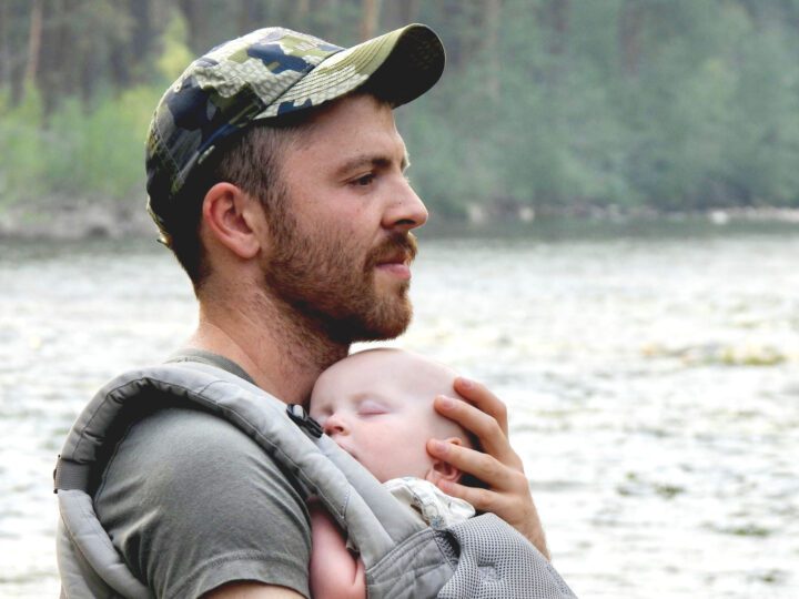 Bearded man with a camouflage cap, holds a sleeping baby in a pouch, against a forest-river background.