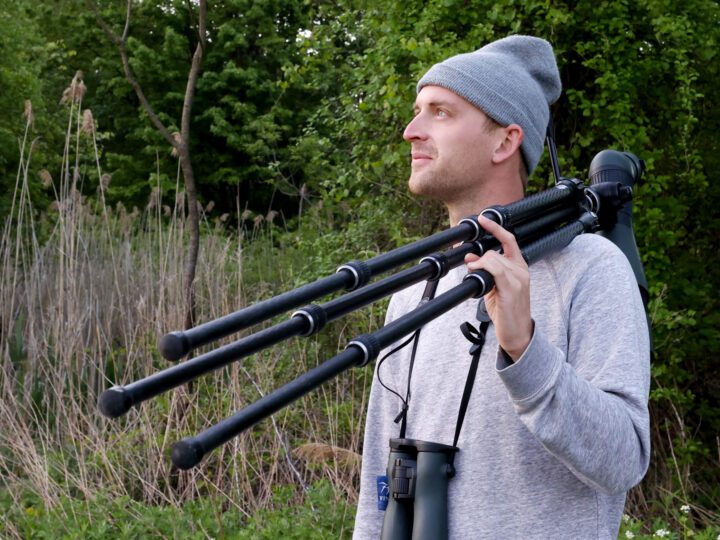 Man in gray cap with large birding scope, stands in the woods.