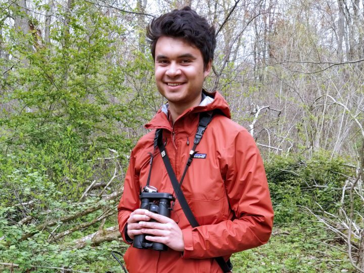 Man with an orange jacket and binoculars stands outdoors.