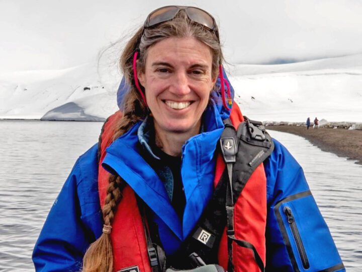 Woman with blue jacket and orange vest with snowy background and lake.