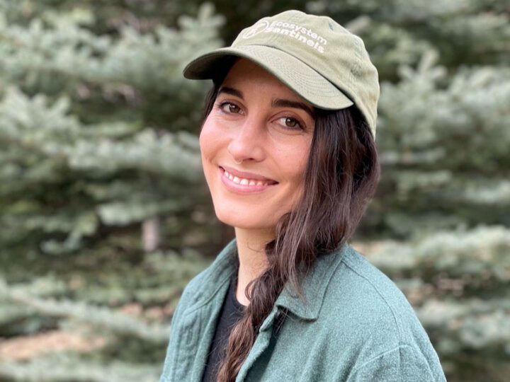 Woman with dark hair and green hat and short, smiles at the camera.