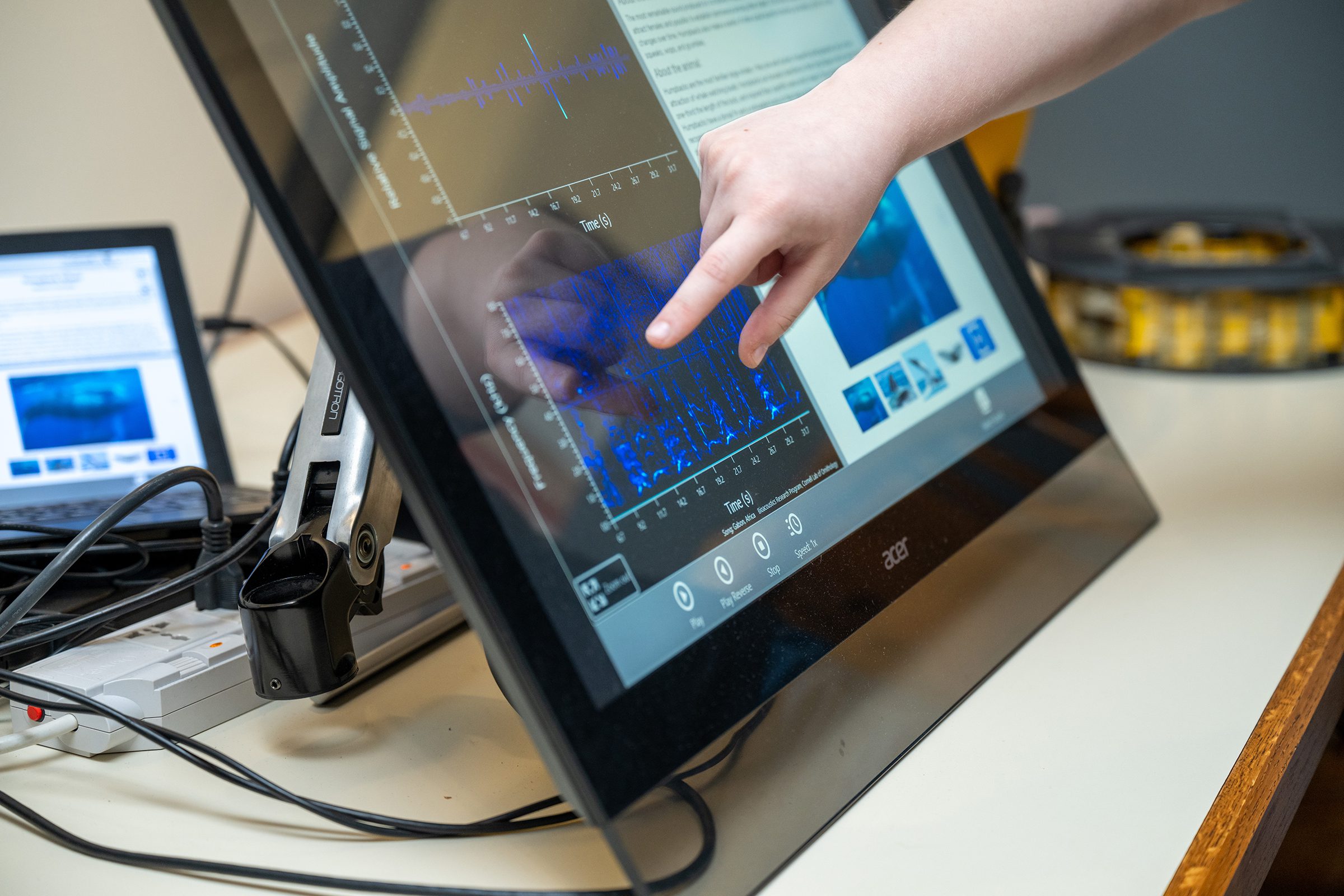 A person’s hand points at a spectrogram on a large touchscreen monitor displaying blue frequency patterns, with a laptop in the background.