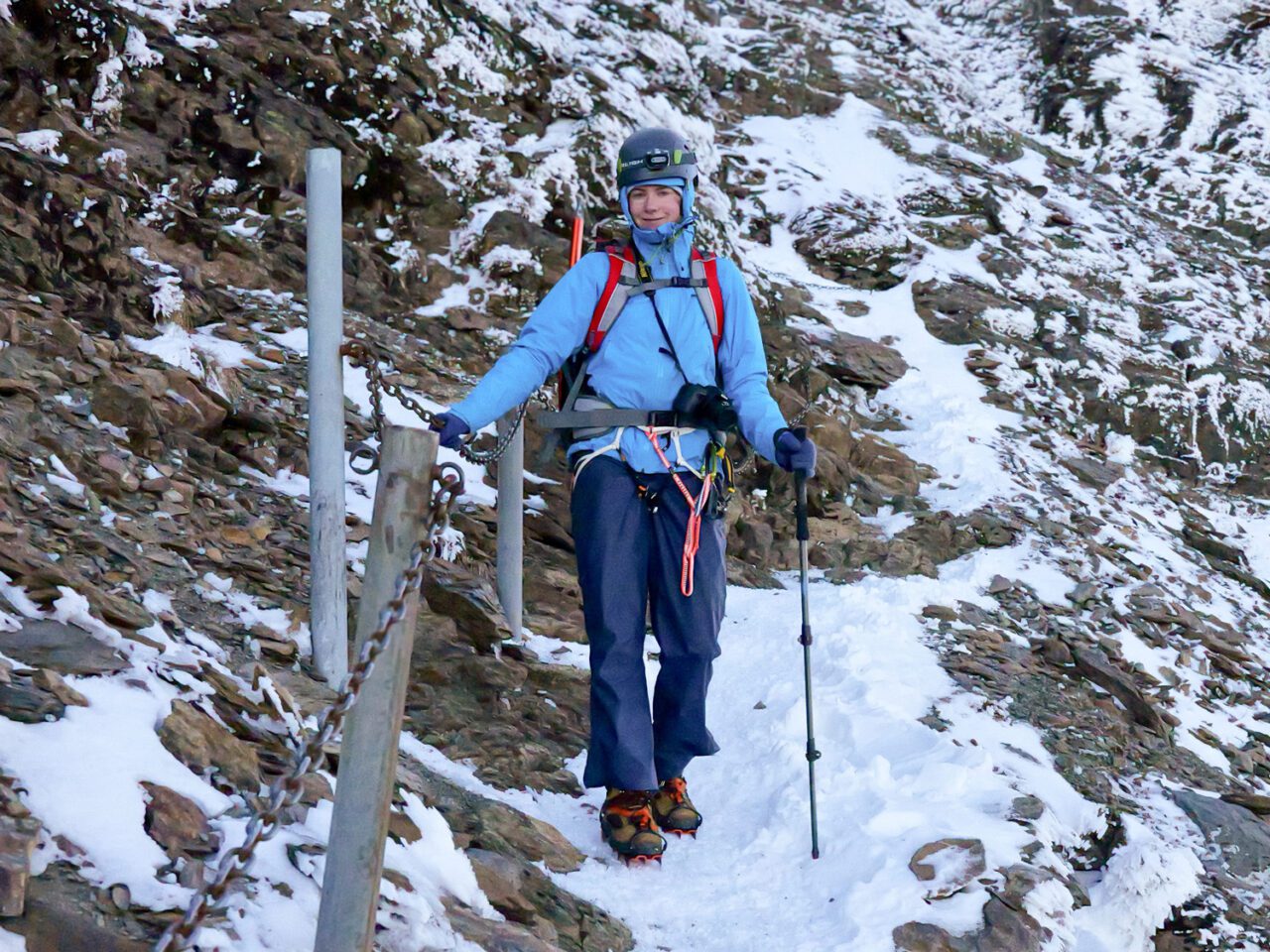 Person wearing climbing gear and helmet walking along a snowy mountain trail
