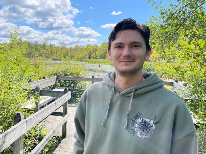 A man in a gray hoodie stands on a wooden boardwalk, looking at the camera.