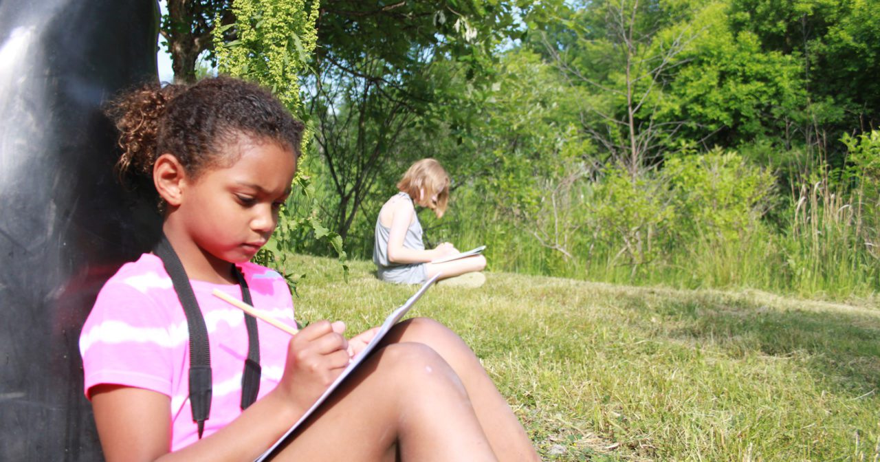 A girl sits outside on a sunny lawn drawing a sound map.