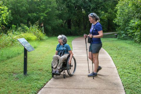 Two people on a concrete trail. One person is in a wheelchair and both are reading the sign.