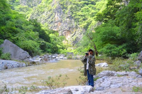 A boy in a camoflague jacket using binoculars next to a river