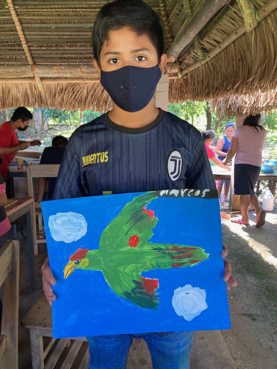 A boy in a facemask holds a painting of a parrot