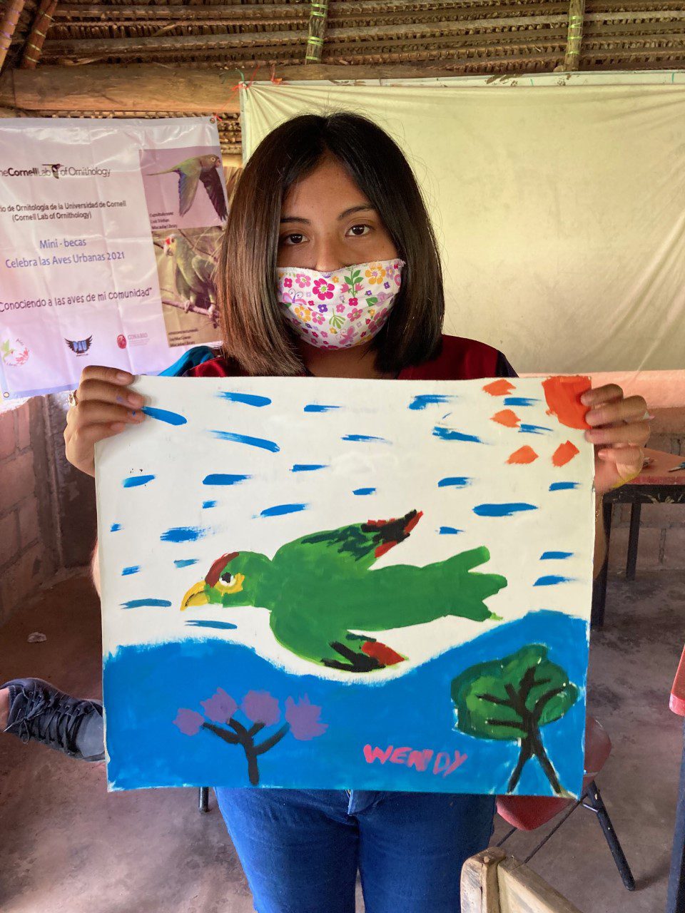 A woman in a floral facemask holds a painting of a parrot