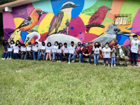 A group of students and teachers sit in front of a bird mural holding certificates