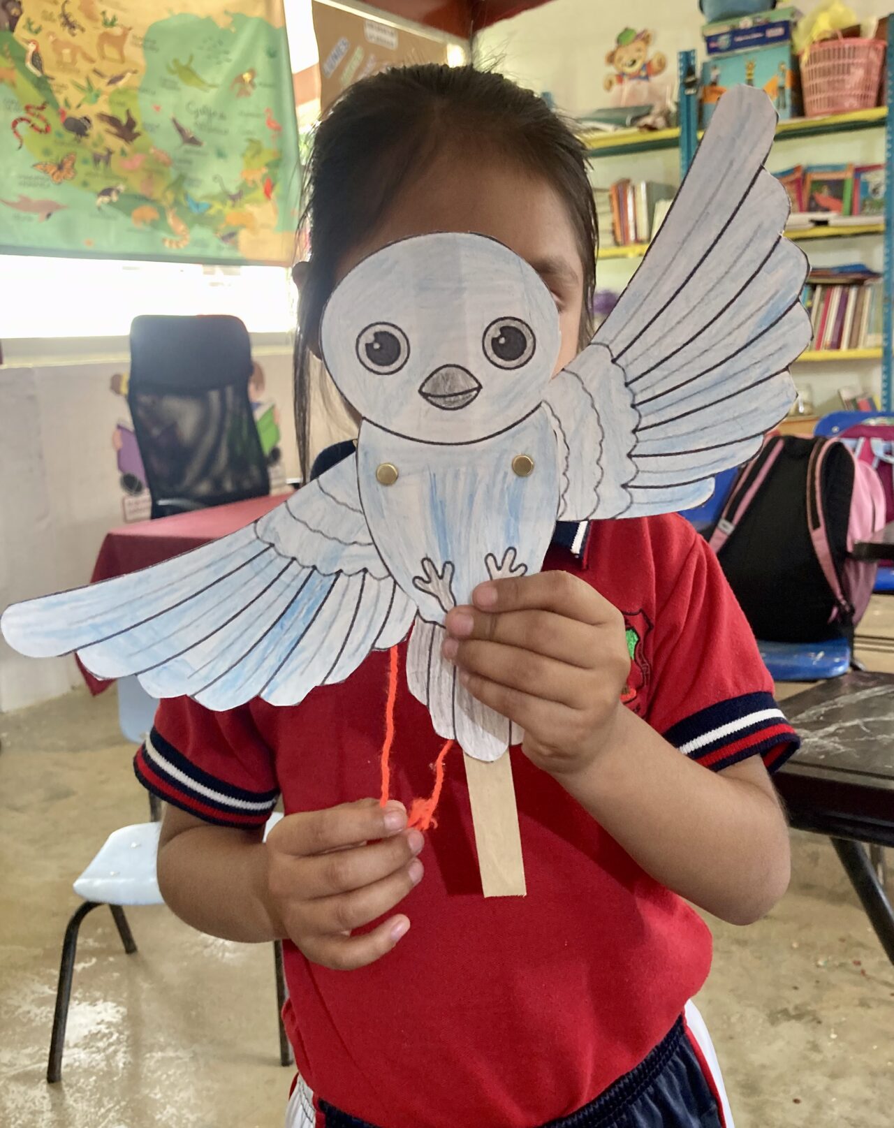 A young student in a red uniform is holding up a handmade paper bird craft in front of their face. The bird is colored light blue and has movable wings attached with brass fasteners. A wooden stick serves as a handle, and orange string represents the bird’s legs.