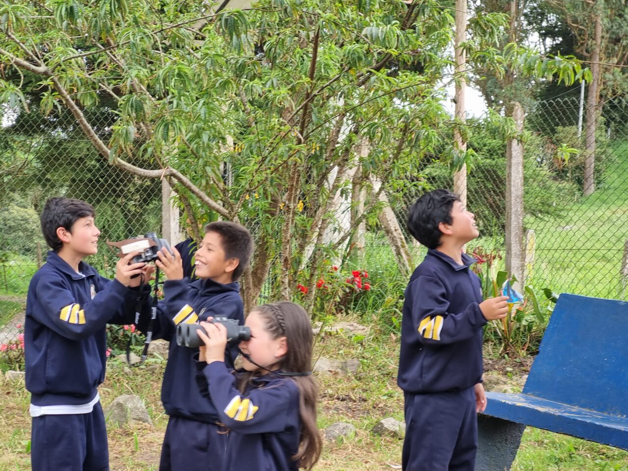 Hay cuatro estudiantes sonriendo en uniforme, mirando a su alrededor. Una niña sostiene un binocular y mira a lo lejos. Dos niños se ríen y comparten unos binoculares.