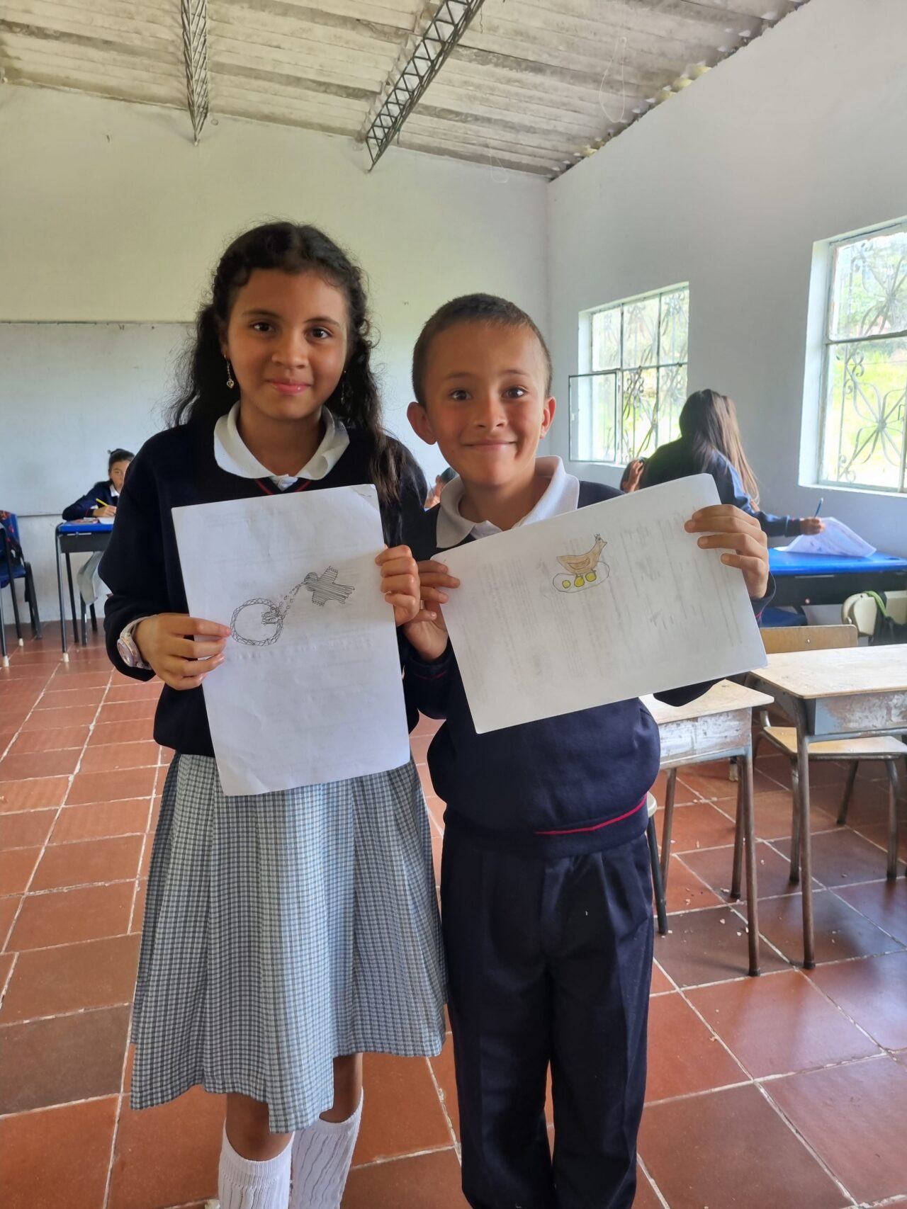 Dos estudiantes sostienen con orgullo sus dibujos ante la cámara. La niña en la izquierda sostiene un dibujo de un pájaro alimentando a sus pollitos. El niño en la derecha sostiene su dibujo de un pájaro sentado sobre sus huevos.