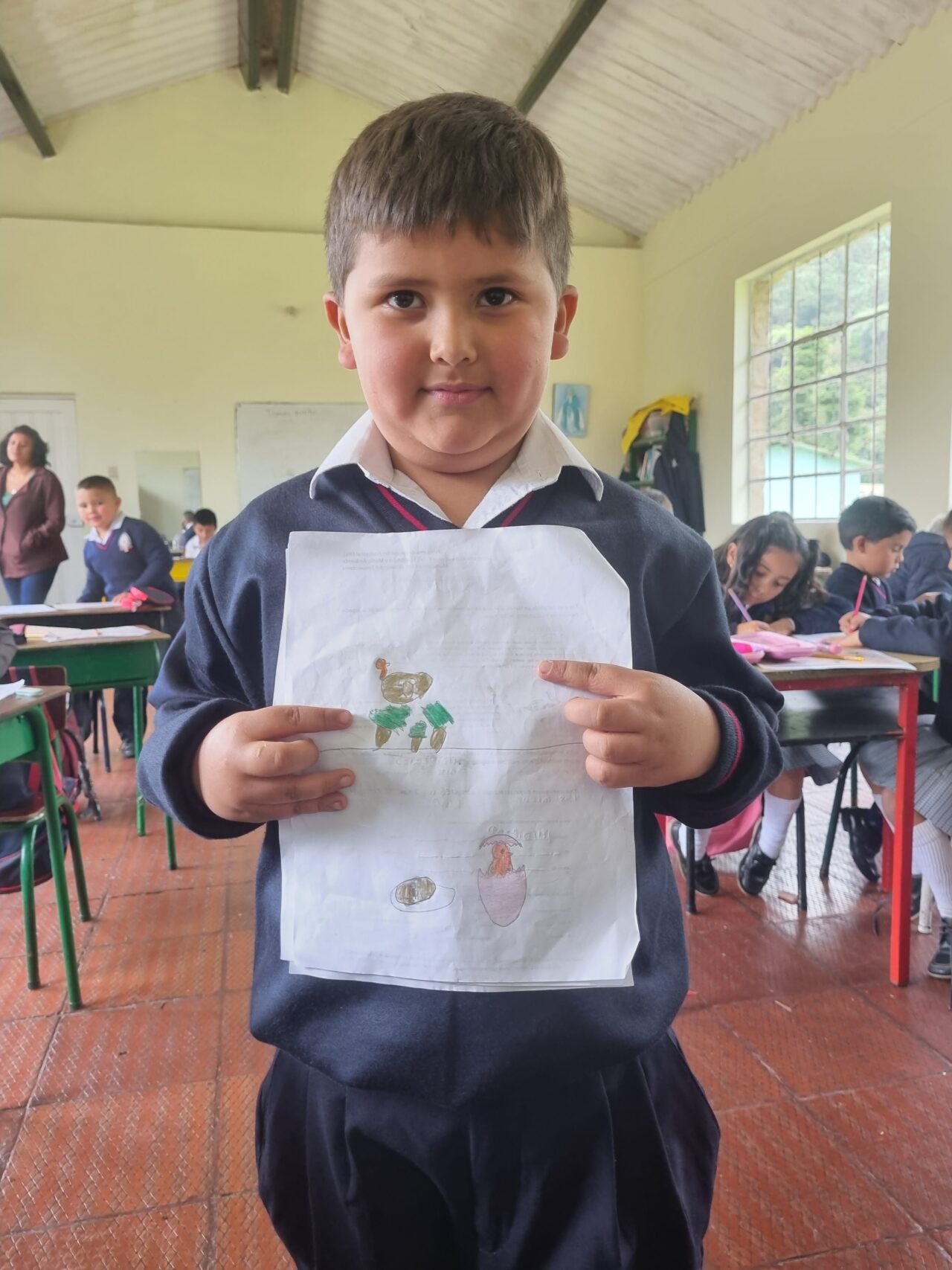 Un niño sostiene con orgullo su dibujo ante la cámara. Su dibujo tiene un pájaro café rodeado por tres árboles y un pájaro anaranjado naciendo de su huevo.
