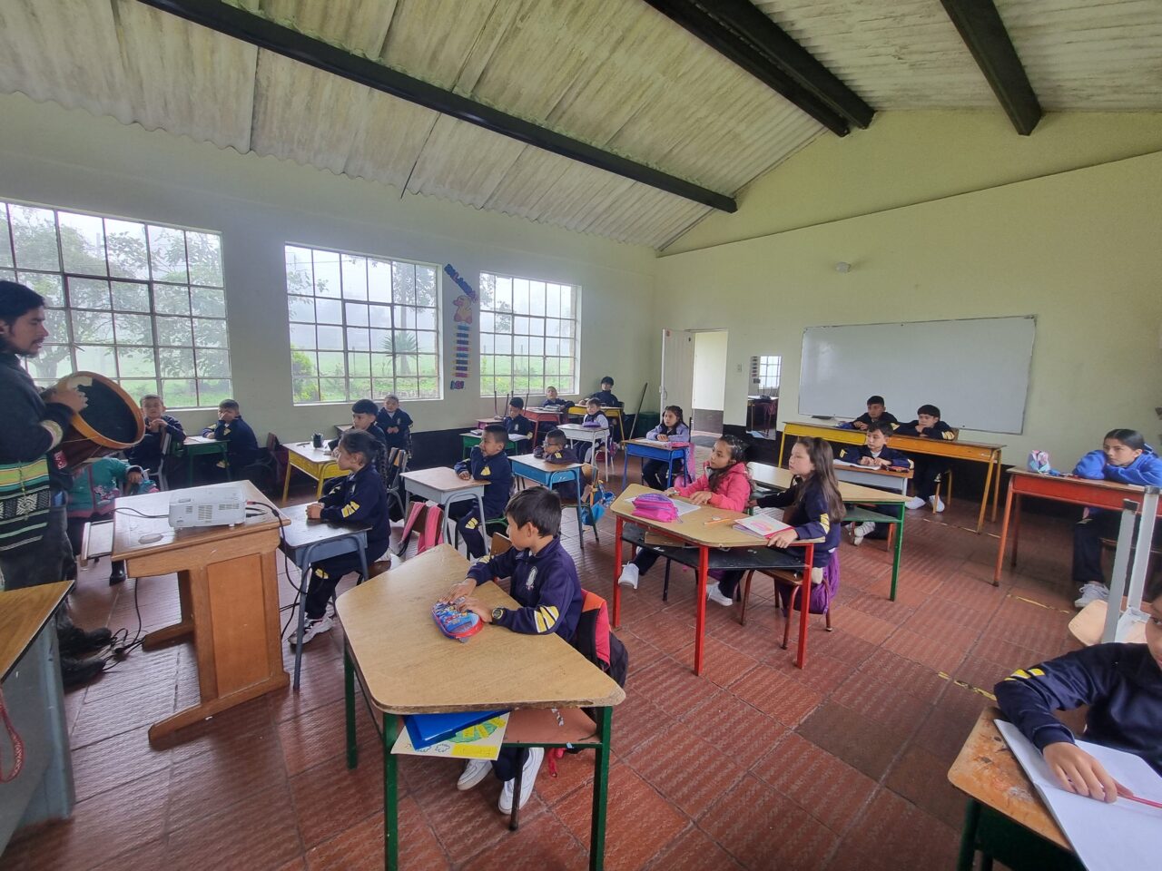 Varios estudiantes están sentados en un salón de clases. Están mirando al frente del salón y están concentrados en su maestro que sostiene un tambor. El maestro les está enseñando a los estudiantes sobre el tambor.