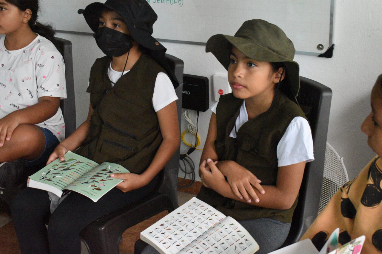 Two children wearing hats and vests sit in a classroom, holding bird field guides and attentively listening to a lesson.