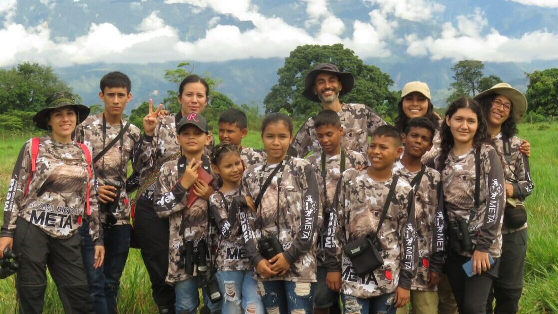 Participants of Global Big Day, including young birders and mentors, pose in a lush green field with mountains in the background. Dressed in matching camouflage shirts, they are equipped with binoculars and cameras, ready to observe and document bird species in their local environment.