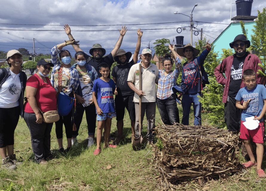 Casa Raizal members gather outdoors, smiling and raising their hands during a community activity. A man holds a shovel, and a composting structure made of sticks and leaves sits in the foreground. The setting includes greenery, a bright blue house, and a partly cloudy sky