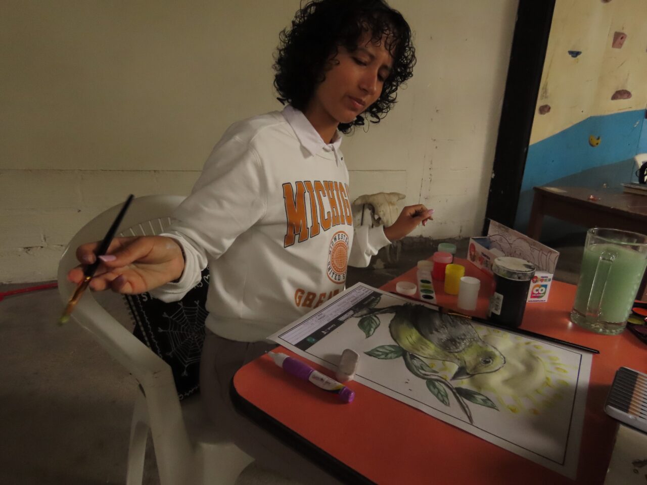 There is a person observing their drawing while holding a paint brush. The painting is a watercolor of a green bird.